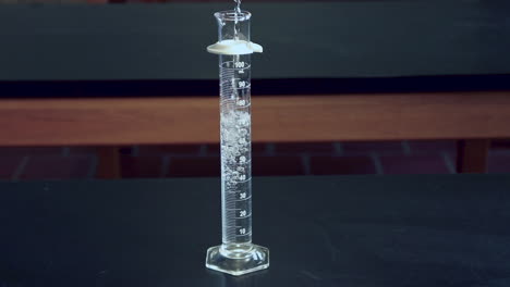 Clear-liquid-being-poured-into-a-100mL-graduated-cylinder-on-a-black-lab-bench-in-a-high-school-chimistry-classroom