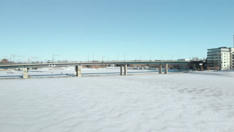Aerial,-drone-shot,-above-Pielisjoki,-towards-traffic-on-Suvantosilta-bridge,-and-apartment-buildings,-on-the-river-shore-on-a-sunny,-winter-day,-in-Joensuu,-North-Karelia,-Finland