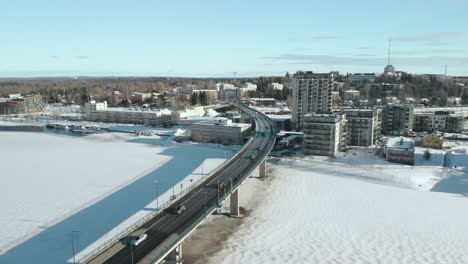 Aerial,-tracking,-drone-shot,-over-Pielisjoki-and-Suvantosilta-bridge,-full-of-cars-and-traffic,-overlooking-apartment-houses-and-buildings,-on-a-sunny,-winter-day,-in-Joensuu,-North-Karelia,-Finland