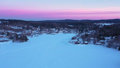 Flying-down-towards-a-small-town-on-the-shore-of-a-frozen-lake-after-dusk-with-a-pink-sky