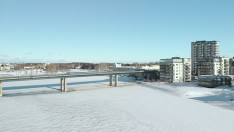 Aerial,-reverse,-drone-shot,-panning-above-Pielisjoki,-away-from-traffic-on-Suvantosilta-bridge,-and-apartment-buildings,-on-the-river-shore-on-a-sunny,-winter-day,-in-Joensuu,-North-Karelia,-Finland