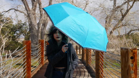 Eine-Hübsche-Frau,-Die-Bei-Schlechtem-Wetter-Und-Einem-Regensturm-In-Zeitlupe-Mit-Einem-Blauen-Regenschirm-über-Eine-Brücke-Geht