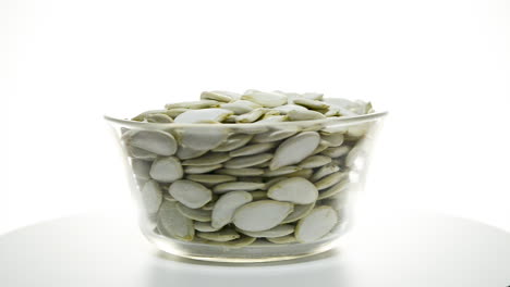 Glass-bowl-full-of-raw-pumpkin-seeds-rotating-on-isolated-white-studio-background