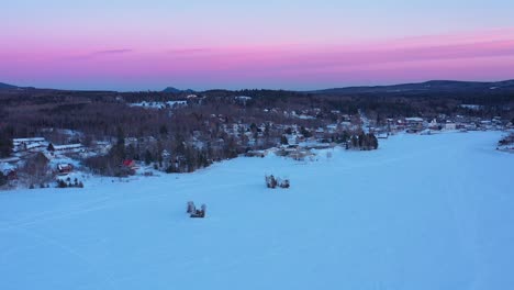 AERIAL-SLIDE-along-the-shore-of-a-frozen-lake-after-dusk-with-a-pink-sky-past-several-camps