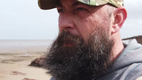 Bearded-urban-man-wearing-baseball-cap-with-mental-health-frustration-looking-across-windy-beach