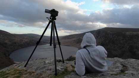 Enthusiastic-photographer-making-time-lapse-at-lake-Guinness-in-Ireland,-sitting-on-rocks-with-hoodie-next-to-hes-camera-on-tripod