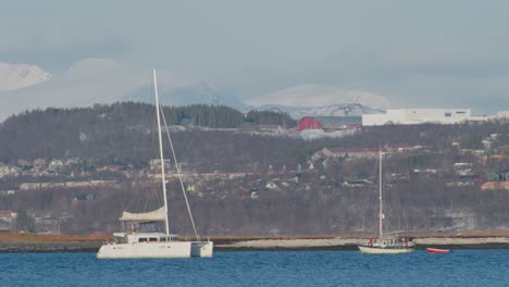 small-white-boat-sailing-along-the-fjord-in-tromso-northern-norway,-telephoto-shot-in-60fps-slow-motion