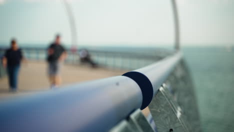 Close-up-of-rail-by-water-leading-out-to-a-pier-as-people-walk-in-slow-motion-in-the-background