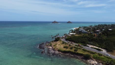 4K-cinematic-drone-shot-of-cars-driving-around-a-bend-near-Kailua-Beach-in-Oahu-contrasted-with-the-blue-ocean-and-two-islands-in-the-background