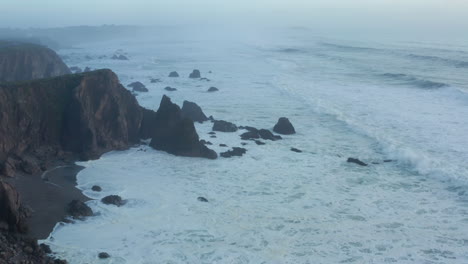 Misty-aerial-seascape,-flying-over-rugged-coastal-cliffs-and-white-ocean-waves-in-fog