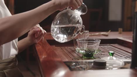 Chinesische-Frau-Gießt-Heißes-Wasser-In-Glasschale-Mit-Chinesischem-Grünem-Tee-Auf-Traditionellem-Teetisch-Aus-Holz