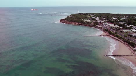 Reverse-aerial-footage-looking-out-over-the-lighthouse-and-Point-Lonsdale,-with-a-cargo-ship-heading-out-into-Bass-Strait,-Victoria