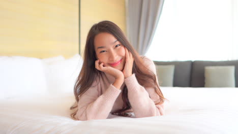 Portrait-of-Young-Smiling-Asian-Woman-Lying-on-Bed,-Leaning-Head-on-Hands-and-Looking-at-Camera,-Slow-Motion