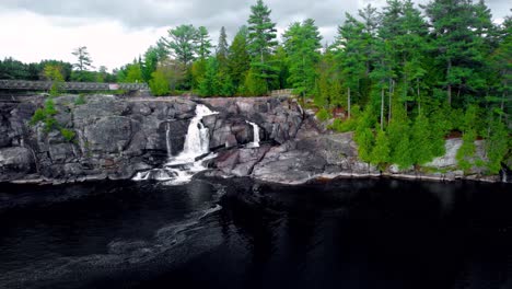 Rocky-waterfall-landscape-fly-over-in-vibrant-northern-Muskoka-with-dark-lake,-gushing-water-High-Falls-lookout-point