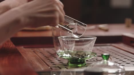 Asian-woman-puts-green-tea-leaves-from-glass-tray-into-Chinese-gaiwan-bowl-on-traditional-wooden-tea-table