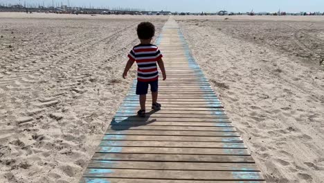 Niño-Africano-De-Dos-Años-Caminando-Sobre-Una-Puerta-De-Madera-En-Una-Playa-De-Arena-Blanca-En-Valencia,-España