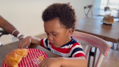 Exotic-and-cute-afro-european-two-year-old-child-wearing-a-eating-a-croissant-and-playing-with-a-plastic-fork,-unfocused-background