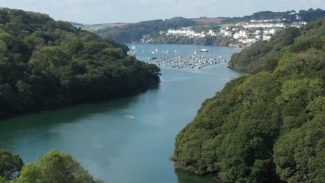 Low-aerial-view-pushing-down-the-River-Fowey-in-Cornwall,-UK-towards-the-coastal-town-of-Fowey-with-paddle-boarders-in-frame