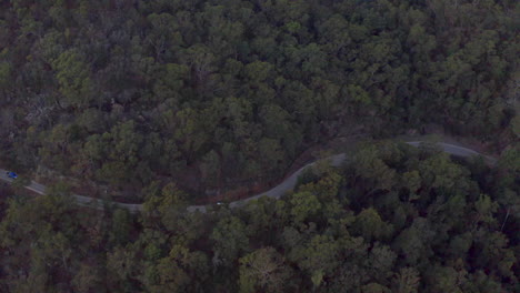 Aerial:-Drone-following-a-white-vehicle-as-it-drives-along-a-winding-road-through-a-forest