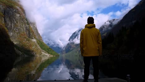 Man-in-a-yellow-raincoat-standing-in-front-of-a-crystal-clear-Fjord-of-towering-mountains