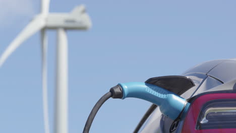 CLOSEUP---An-unbranded-electric-car-charges-in-front-of-a-wind-turbine,-sunny-day