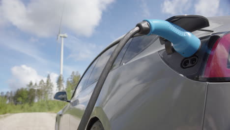 WIDE-SHOT---A-generic-electric-car-charges-in-front-of-a-wind-turbine