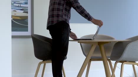 UK-Asian-Male-Walking-Towards-Table-Carrying-Laptop-And-Logging-On-In-Remote-Working-Space