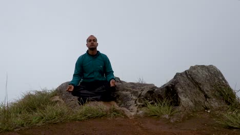 Hombre-Meditando-En-La-Roca-De-La-Montaña-Con-Fondo-De-Niebla-Blanca-Desde-Un-ángulo-Plano
