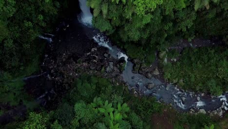 A-drone-shot-of-wild-flowing-creek-passing-fast-grey-ball-shaped-stones-and-green-grass-blades-close-to-the-waterfall-Kedung-Kayang,-Magelang,-East-Java,-Indonesia