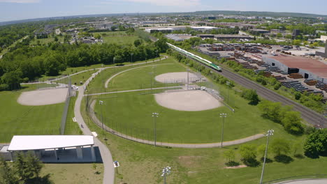 Aerial-view-of-commuter-train-passes-by-an-empty-baseball-diamond-on-a-sunny-summer-day