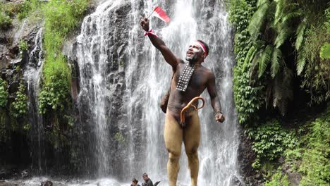 Papua-man-wearing-traditional-clothes-of-Dani-tribe,-red-white-headband-and-bangle-is-holding-little-Indonesia-flag-and-celebrating-Indonesia-independence-day-against-waterfall-background