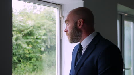A-sad-pensive-lonely-businessman-looking-up-at-a-grey-sky-and-rain-outside-of-his-office-window