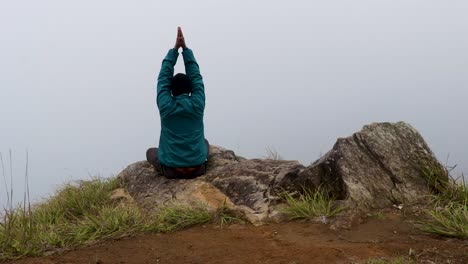 Hombre-Meditando-En-La-Roca-De-La-Montaña-Con-Fondo-De-Niebla-Blanca-Desde-Un-ángulo-Plano
