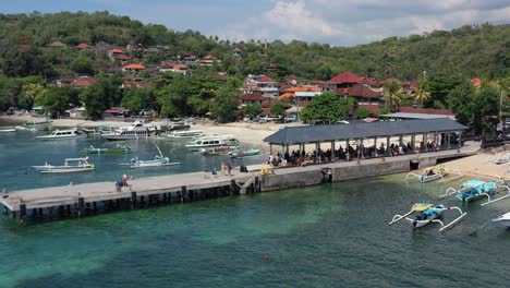 Touristen-Auf-Einem-Pier-Im-Hafen-Von-Padangbai-Warten-Auf-Eine-Fähre-Von-Bali-Indonesien-Mit-Weißem-Sandstrand-Und-Fischerbooten,-Die-Im-Türkisblauen-Wasser-Verankert-Sind,-Antenne
