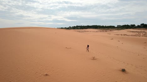 female-walking-up-a-red-desert-sand-dune-landscape-on-a-cloudy-day-in-Mui-Ne-Vietnam
