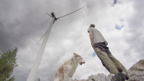 One-man-and-his-dog-stand-below-a-huge-wind-turbine,-before-walking-away