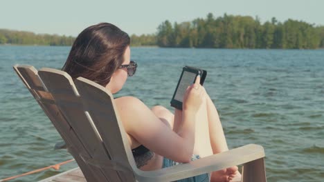 Camera-pans-over-a-woman-relaxing-and-reading-on-a-dock-next-to-a-lake-in-the-summer-season