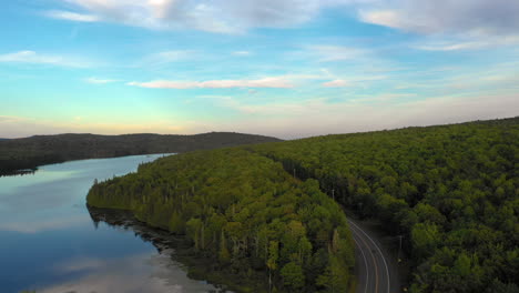 Aerial-view-of-the-sunset-with-a-road-going-through-a-dense-forest-with-a-mirror-like-pound-on-the-side