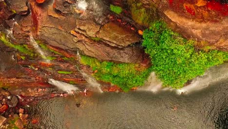 Birds-eye-drone-shot-flying-along-the-edge-of-a-waterfall-cliff-as-people-swim-below