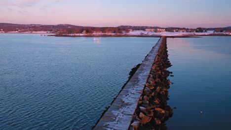 Aerial-footage-flying-diagonally-over-a-snow-dusted-rocky-breakwater-in-Rockland-Maine-at-sunrise