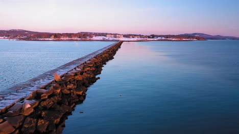 Aerial-footage-flying-towards-shore-along-side-of-a-snow-dusted-rocky-breakwater-in-Rockland-Maine-at-sunrise
