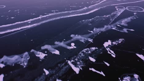 Close-up-Aerial-TOP-DOWN-view-of-the-dark-ice-of-a-frozen-lake-with-small-islands-of-snow-that-transitions-over-to-a-lighter-thicker-ice