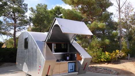 A-new-deluxe-metal-teardrop-travel-trailer-in-a-forest-campsite-with-its-back-open-to-the-kitchen-and-stove-area-REVEAL