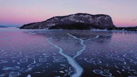 Flying-low-and-slow-at-dawn-over-the-cracks-in-a-frozen-lake-towards-the-cliff-face-of-an-isolated-mountain