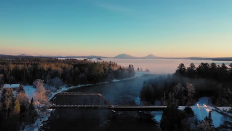 Flying-over-the-mist-of-a-curving-winter-river-past-a-railroad-trestle-towards-a-snow-covered-frosted-lake-with-two-prominent-mountains-in-the-distance