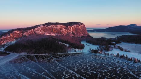 Aerial-PULL-BACK-and-LIFT-UP-away-from-the-peak-of-a-cliff-faced-mountain-during-a-winter-sunrise-revealing-the-misty-lake-and-endless-forests-behind