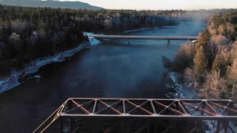 Luftaufnahmen-Fliegen-Langsam-über-Einen-Eisenbahnbock-Und-In-Richtung-Einer-Brücke,-Die-Bei-Sonnenaufgang-Einen-Winterlichen-Fluss-überquert