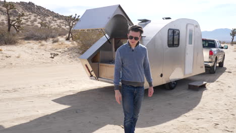 Slow-motion-shot-of-a-handsome-white-man-on-a-desert-camping-trip-next-to-a-metal-travel-trailer-tiny-house