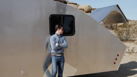 A-handsome-young-white-man-traveler-leaning-against-a-metal-teardrop-travel-trailer-tiny-house-on-his-solo-road-trip