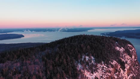 Aerial-view-flying-away-from-the-peak-of-a-cliff-faced-mountain-while-seeing-the-misty-freezing-lake-behind-it-during-a-winter-sunrise